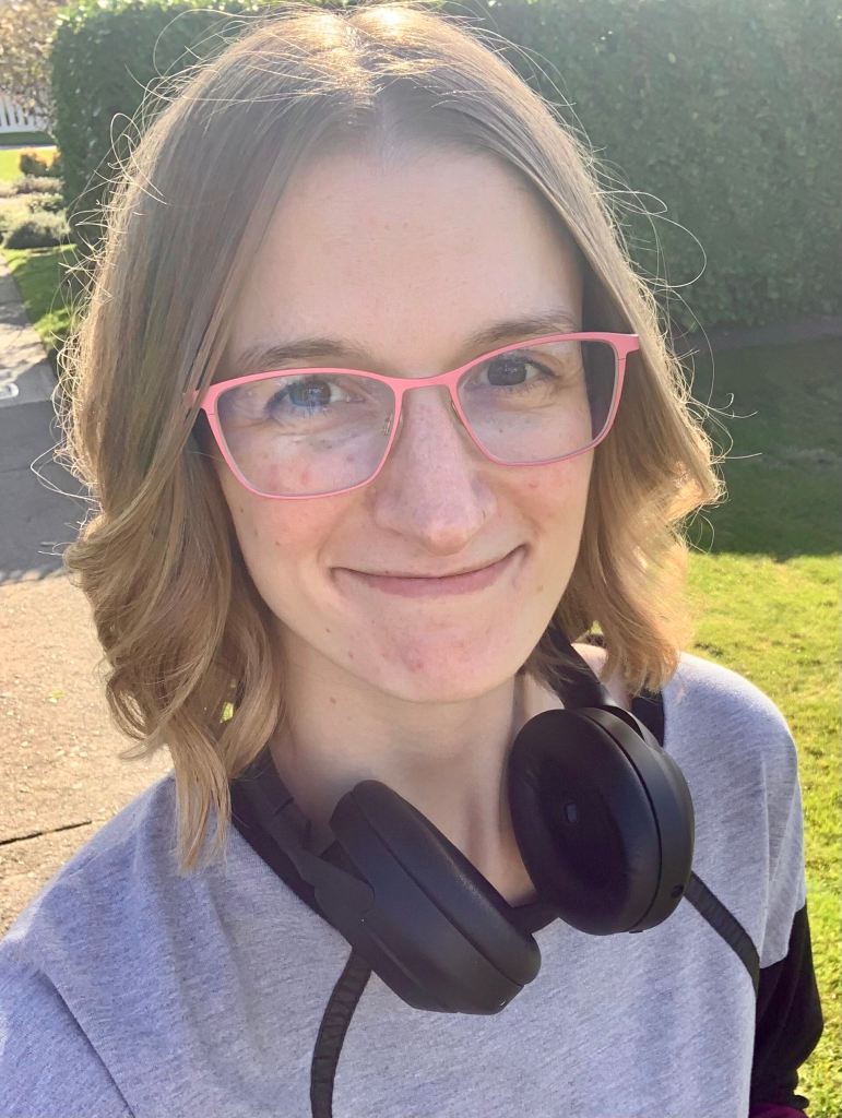 A blonde white woman smiling at the camera wearing a pair pink-framed glasses, with large over-ear headphones hanging around her neck